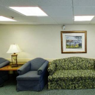 A waiting room with a floral sofa, two blue chairs, a lamp on a wooden table, and a framed painting on the wall.