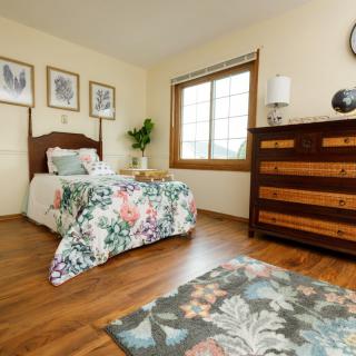 A cozy bedroom featuring floral bedding and wooden furniture, beautifully lit by natural light from a large window.
