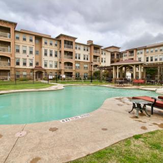 A residential apartment complex with a swimming pool and patio furniture under a cloudy sky.