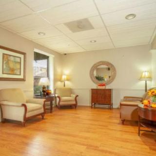 A cozy waiting room featuring comfortable chairs, a table with a lamp, artwork, and a mirror on the wall over a wooden floor.