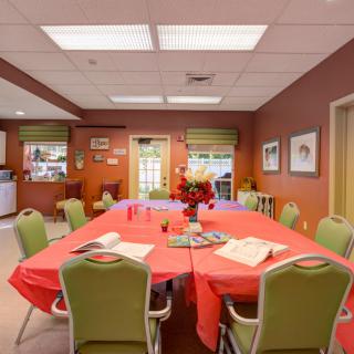 A cozy dining room with a large table covered in a red tablecloth, surrounded by green chairs and decorated with flowers.