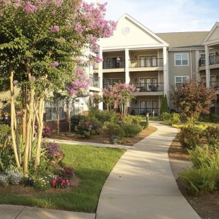 A well-landscaped pathway leading to an apartment building, surrounded by vibrant gardens and trees.