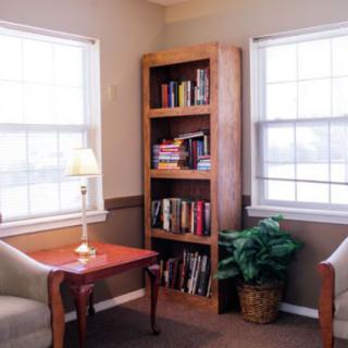 A cozy reading nook with a bookshelf, armchairs, and a warm lamp between two bright windows.