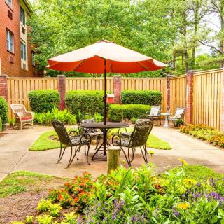 A cozy outdoor patio area with a table, chairs, and an umbrella surrounded by lush greenery.