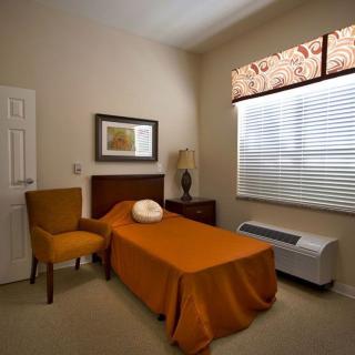 A minimalist bedroom featuring a single bed with an orange bedspread, a chair, and natural light through the window.