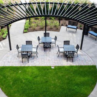 An outdoor patio setup with tables and chairs under a pergola, surrounded by greenery.