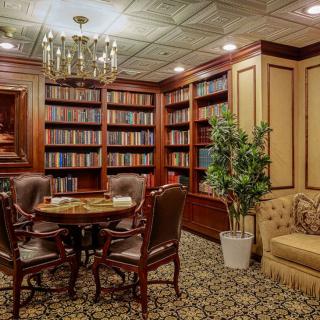 A cozy library room featuring wooden bookshelves, a round table with leather chairs, and a plush sofa under a chandelier.