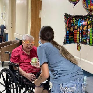 A heartfelt moment of celebration between a caregiver and an elderly individual in a wheelchair, surrounded by festive balloons.