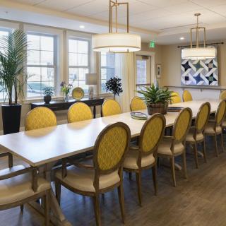 A spacious dining room with a long table surrounded by yellow upholstered chairs, featuring modern lighting and decorative plants.