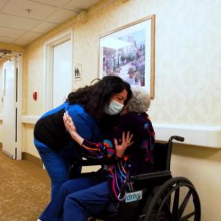 A caregiver wearing a mask hugs an elderly person in a wheelchair in a nursing home hallway.