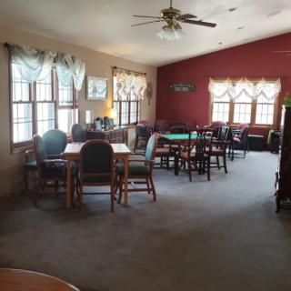 A spacious room with multiple tables and chairs, featuring large windows with curtains and a ceiling fan.