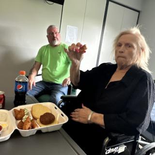 A woman holding food with a man seated in the background, enjoying a meal together.