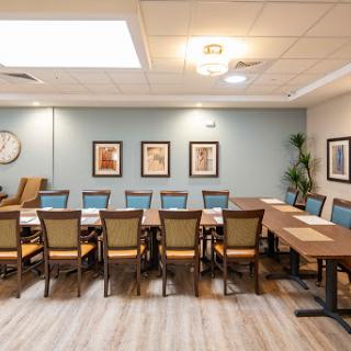 A well-lit conference room featuring a long wooden table surrounded by chairs, complemented by artwork on the walls and natural lighting from a skylight.