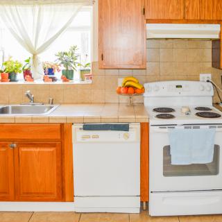 A bright kitchen with wooden cabinets, featuring a stove, oven, dishwasher, and a sink next to a window adorned with plants.