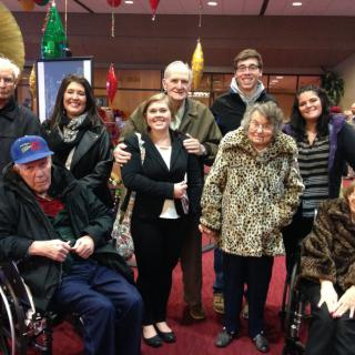A group of people, including elderly individuals and some in wheelchairs, posing together indoors with smiling faces.