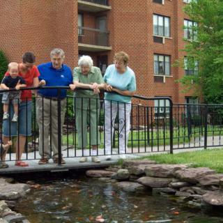 A family group admires a pond from a small bridge in an outdoor setting near an apartment building.