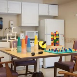 A therapy room featuring colorful stacking cups and building blocks on a table, with a kitchenette in the background.