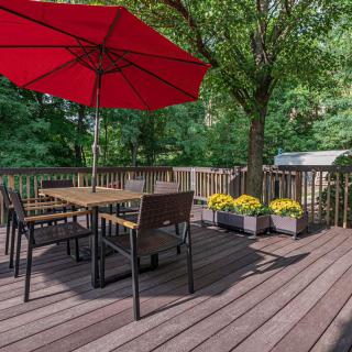 A serene outdoor patio with a wooden deck, featuring a dining table set under a red umbrella, surrounded by lush trees and potted flowers.