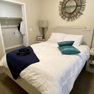 A cozy bedroom with a neatly made bed, featuring teal pillows and a navy throw blanket. A decorative mirror hangs above the headboard, with a lamp and nightstand nearby.
