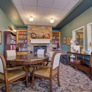 A cozy library with a fireplace, surrounded by bookshelves and a wooden table with chairs.