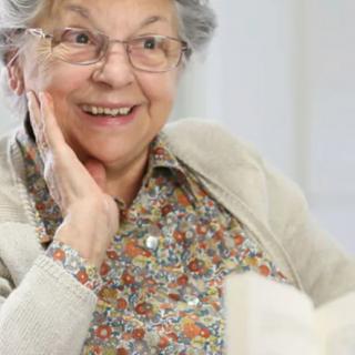 An elderly woman smiling warmly while reading a book indoors.