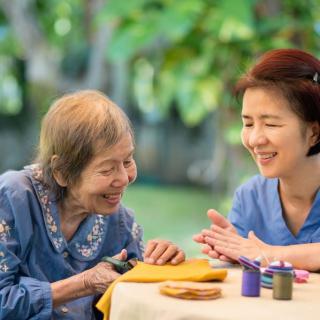 An elderly woman and a younger woman enjoy sewing together outdoors, sharing smiles and companionship.