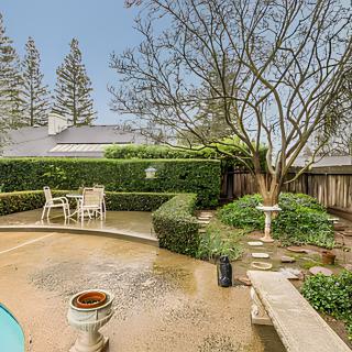 A serene backyard patio area with outdoor seating, surrounded by lush hedges and a tree.
