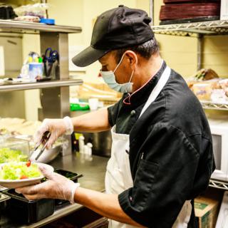 A chef wearing a mask and gloves prepares a dish in a restaurant kitchen.