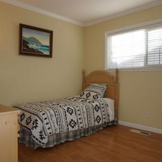 A minimalist bedroom featuring a single bed with patterned bedding, a painting on the wall, and natural light streaming through a window.