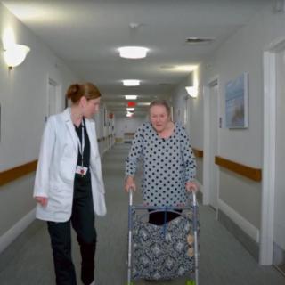 A doctor walks alongside an elderly patient using a walker in a hospital hallway.