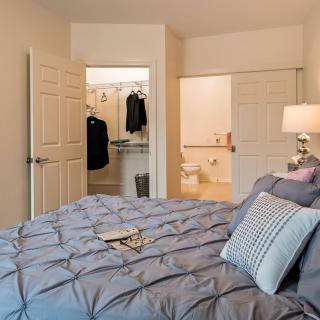 A cozy bedroom featuring a neatly made bed with pillows, an open closet, and a bedside lamp.