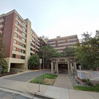 A view of a multi-story building with an entrance surrounded by trees and a sidewalk.