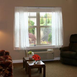 A cozy living room with a window adorned with sheer curtains, featuring an armchair, sofa, and a small table.