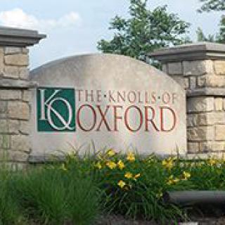 A stone sign for 'The Knolls of Oxford' with surrounding greenery and yellow flowers.