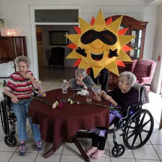 Elderly individuals sitting around a table, joined by someone in a sun costume.
