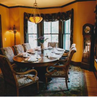 An elegant dining room with a set dining table, chandelier, and grandfather clock by the bay windows.
