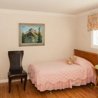 A cozy bedroom with a single bed dressed in pink, a teddy bear on the bed, and a painting hanging above. A dark chair sits next to the bed near a window with curtains.