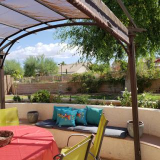 A cozy outdoor patio featuring a colorful table and comfortable seating under a sunshade, surrounded by greenery.