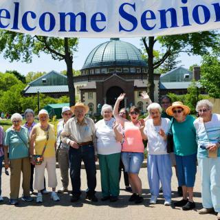 A group of seniors gathers under a 'Welcome Seniors' banner at an outdoor event.