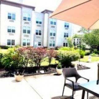 A sunny patio area with chairs and a table under an umbrella, overlooking a garden and modern building.