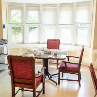 A cozy dining area featuring a round table surrounded by red upholstered chairs, illuminated by natural light from large windows.