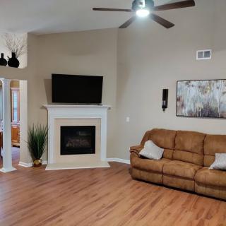 A cozy living room featuring a brown sofa, a fireplace with a TV mounted above it, and wooden flooring. The space is decorated with artwork and houseplants.