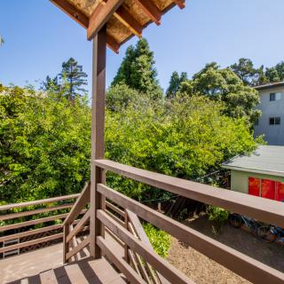 A sunny view from a wooden balcony overlooking lush green trees and a neighboring house.