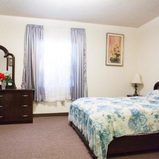 A cozy bedroom featuring a bed with floral bedding, a wooden dresser with a mirror, and curtains framing the window.