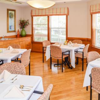 A cozy dining room with neatly arranged tables and chairs, featuring a wooden floor and natural lighting from large windows.