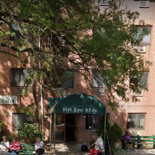 People seated on benches outside a building entrance surrounded by trees.