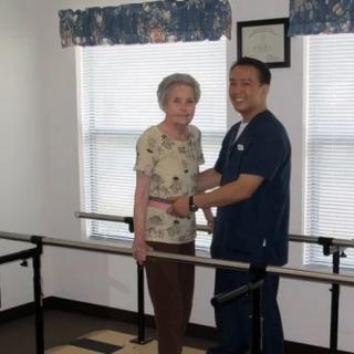 An elderly woman receives assistance from a caregiver during physical therapy.