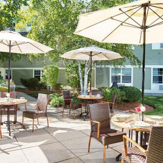 A sunny outdoor patio with tables and chairs under large umbrellas, surrounded by greenery.