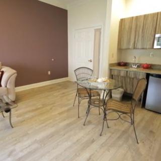 A modern living space featuring a dining area with a glass table and metal chairs, adjacent to a compact kitchenette with wood finish cabinets and light wood flooring.