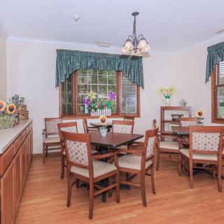 A cozy dining room with wooden flooring, featuring a central table surrounded by chairs and adorned with floral decorations and window curtains.